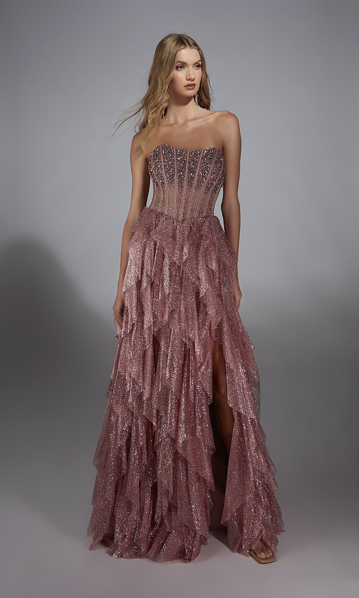 A woman wears the Alyce 61762 Strapless Long Glitter A-Prom Dress featuring ruffles and a sheer, beaded bodice, standing against a plain gray background.
